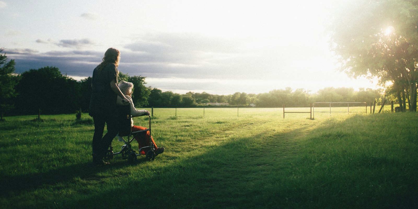 A family caregiver stands beside her grandmother, who is seated in a wheelchair at home